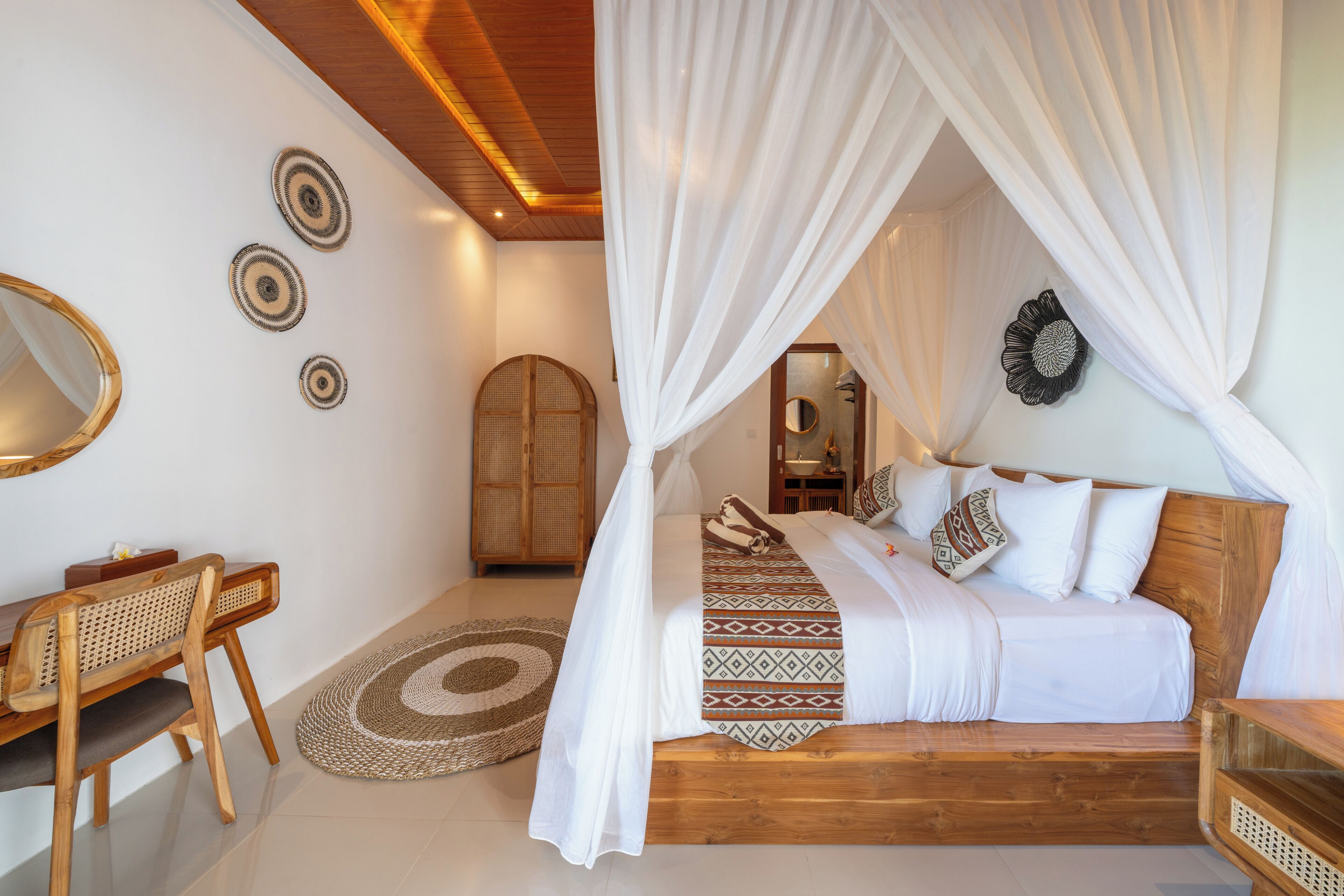 A bedroom with a canopy bed, a wooden desk, a rattan chair, a round woven rug, and a mirror on the wall.