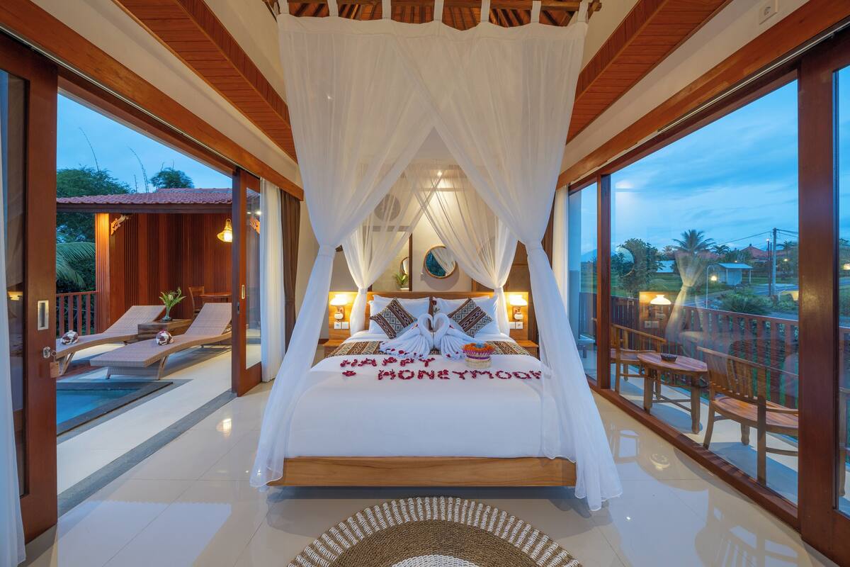 Un dormitorio amplio con una cama grande, vista a la piscina y un balcón con mobiliario.