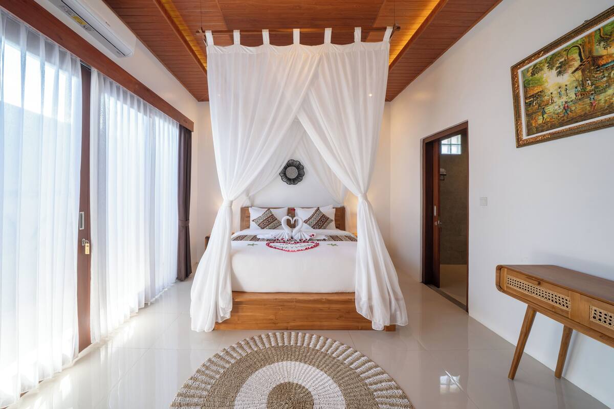 Un dormitorio con una cama con dosel, suelo de madera, un banco de ratán y un cuadro en la pared.