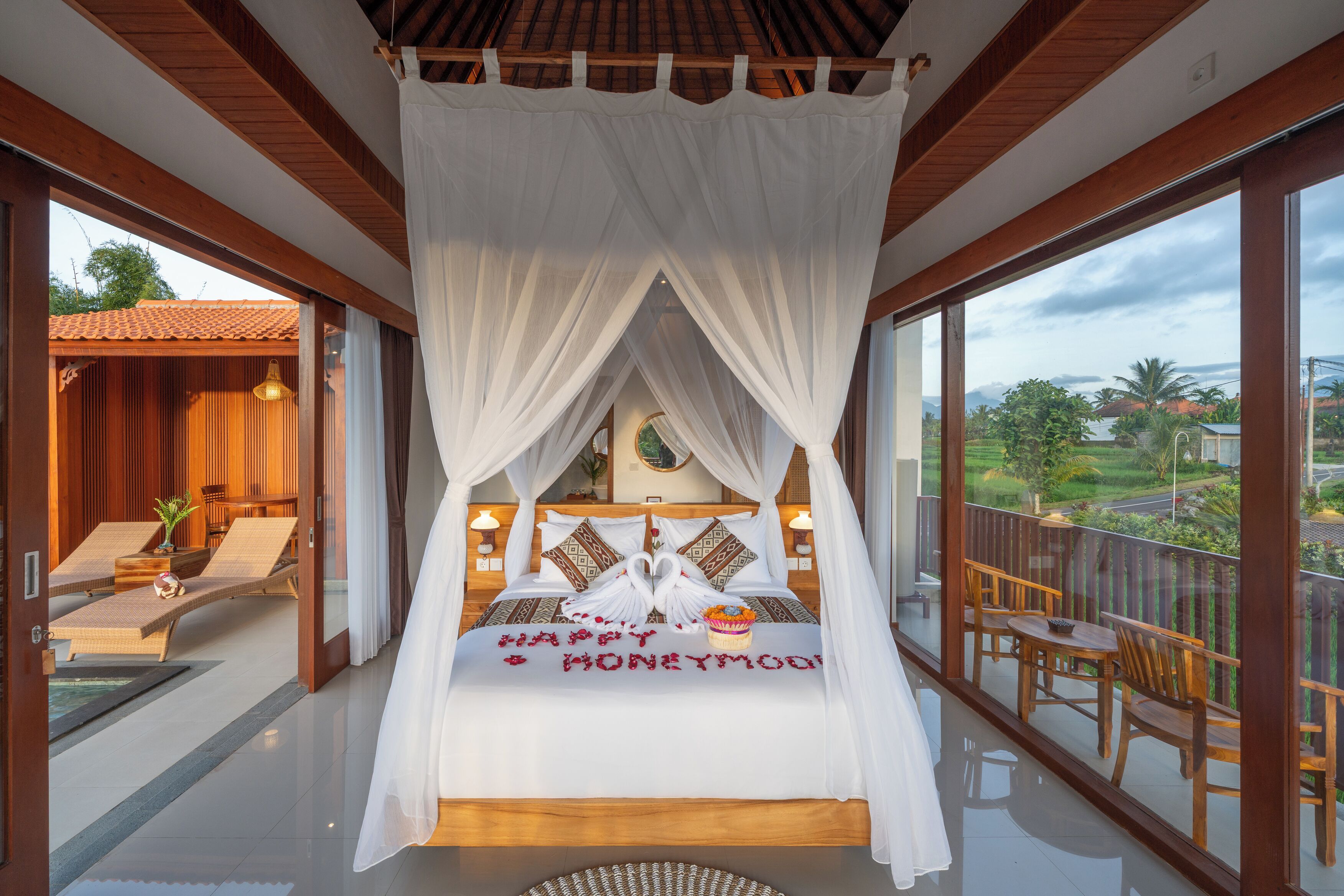 A bedroom with a canopy bed, a balcony with wooden furniture, and a view of the landscape.