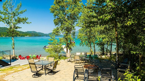 A lakeside patio with outdoor seating, a glass table, and a view of the lake and mountains.