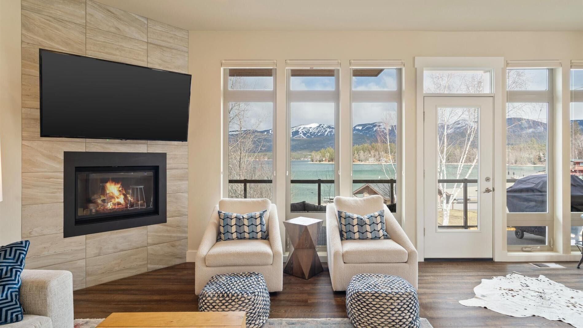 A living room with a fireplace, two armchairs, a coffee table, and large windows with a mountain view.