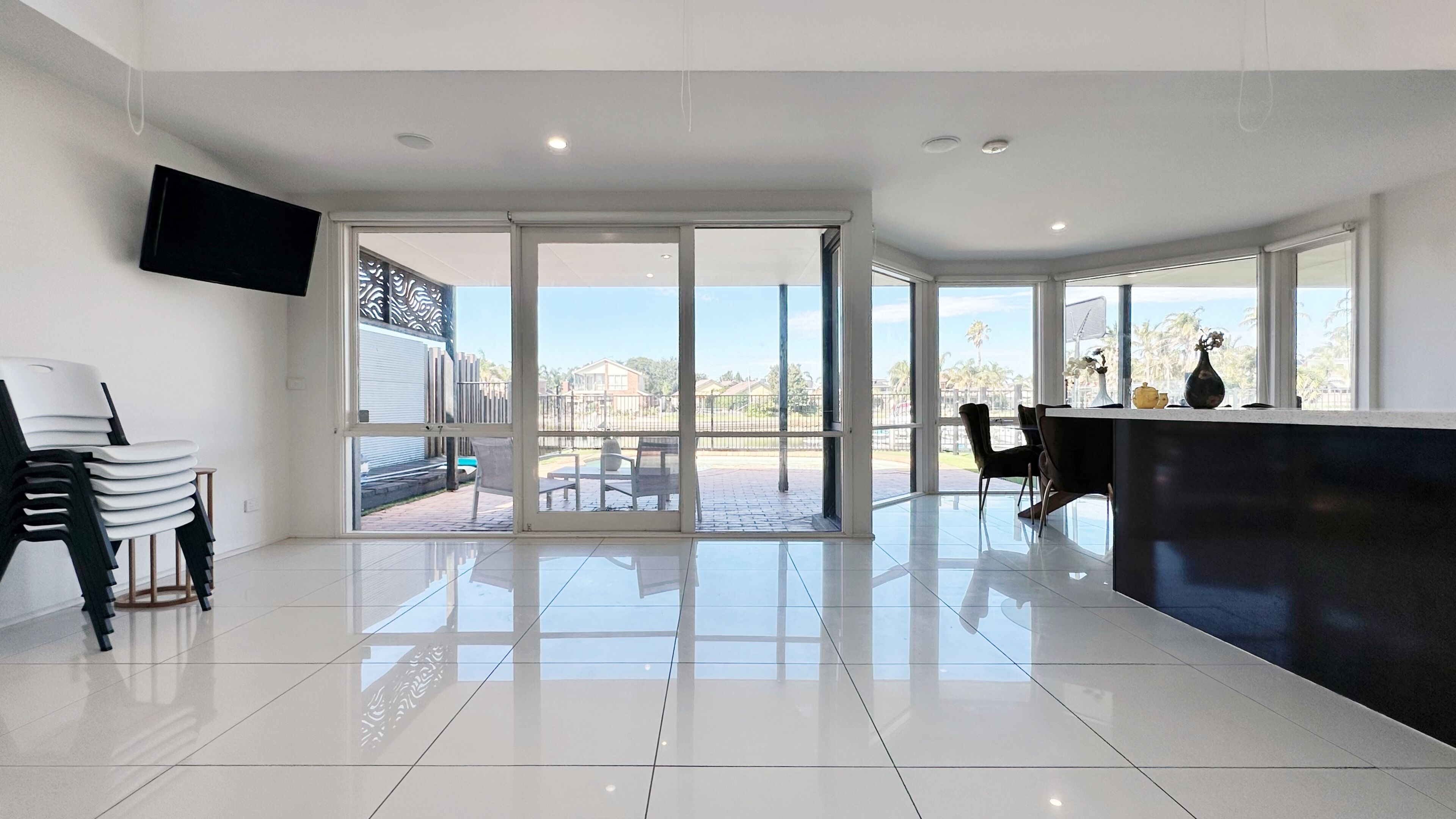 A spacious room with a large sliding glass door leading to an outdoor patio, a flat-screen TV mounted on the wall, and a modern kitchen area with a dark countertop.