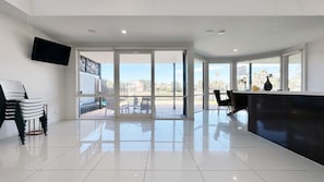 A spacious room with a large sliding glass door leading to an outdoor patio, a flat-screen TV mounted on the wall, and a modern kitchen area with a dark countertop. - Lakefront/View/Swimming Pool/6B4B/Shopping Nearby (Patterson Lakes)