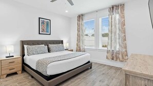 Une chambre à coucher comprenant un lit, des tables de chevet, un ventilateur de plafond, une fenêtre avec des rideaux et un tableau encadré accroché au mur.