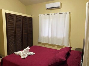 Une chambre à coucher comprenant un lit, une fenêtre avec des rideaux, un climatiseur fixé au mur et une armoire avec des portes à persiennes.