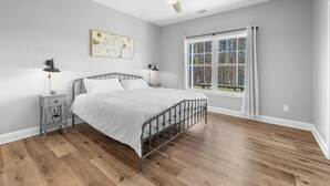 Een slaapkamer met een bed, nachtkastje, plafondventilator, raam met gordijnen en een wandkunstwerk.