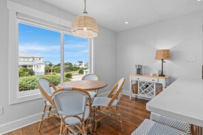 A dining area with wicker chairs, a round table, a hanging light fixture, and a view of houses through large windows. - Welcome to Selah by the Sea, your perfectly located Bald Head Island escape (Bald Head Island)