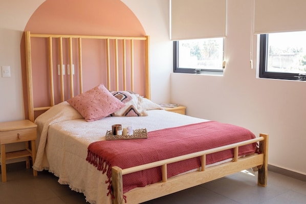 A bed with a wooden frame, a red blanket, and pillows, a bedside table, and a window with a view.