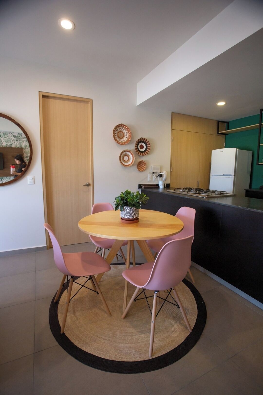 A dining area with a round table, four chairs, a potted plant, and a kitchen in the background.