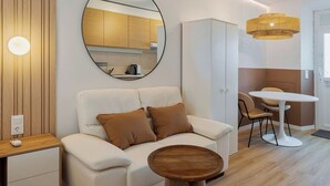 Un salon moderne avec un canapé, une table basse ronde, un coin repas avec une table et des chaises, une cuisine visible dans le miroir et un lustre.