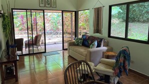 Sala de estar com sofá, cadeiras, mesa de jantar e vista para um jardim através de grandes janelas.