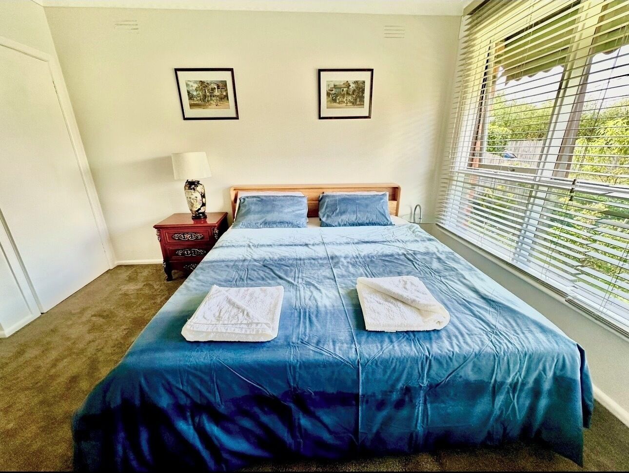 Une chambre à coucher avec un lit, deux tableaux encadrés au mur, une table de chevet et une fenêtre avec des stores.