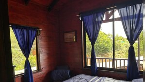Ein Schlafzimmer einer Holzhütte mit einem Bett, zwei mit Vorhängen versehenen Fenstern, einem Stuhl und einer Holzterrasse davor.