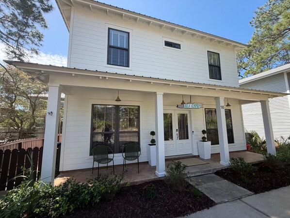 A two-story white house with a covered porch, a sign reading 'SEA GYPSY,' and two potted plants on the porch. - Sea Gypsy - 4BR, Walk to Beach and iconic eateries, Pet Friendly, Hot Tub, Bikes (Santa Rosa Beach)