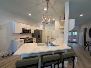A modern kitchen with white cabinetry, stainless steel appliances, a central island with bar stools, and a chandelier. - Sea Gypsy - 4BR, Walk to Beach and iconic eateries, Pet Friendly, Hot Tub, Bikes (Santa Rosa Beach)