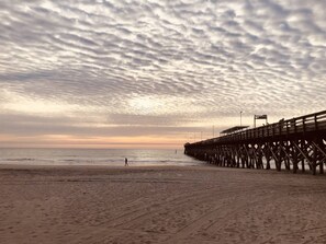 A beach with a wooden pier extending into the ocean, a person walking on the sand, and a cloudy sky. - Stunning Oceanfront King Suite at Oceans One Resort 9th Floor (Myrtle Beach)