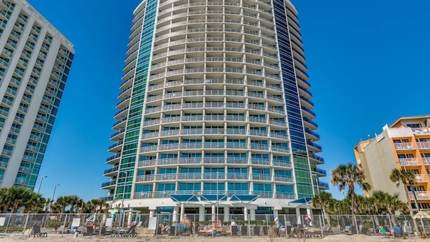 A multi-story hotel with a curved facade, balconies, and a pool area with sun loungers. - Stunning Oceanfront King Suite at Oceans One Resort 9th Floor (Myrtle Beach)