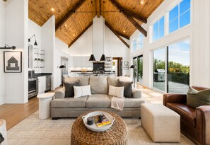 A spacious living area with a high ceiling, a grey sofa, a brown leather sofa, a wicker coffee table, and a kitchen in the background. - Discover Pool Paradise w/ Breathtaking Lake Views • HotTub • Fun Games • Privacy (Blue Ridge)