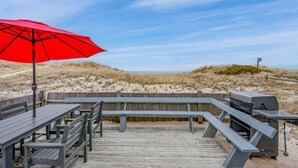 Een houten terras met banken en een barbecue, met uitzicht op een strand met duinen.