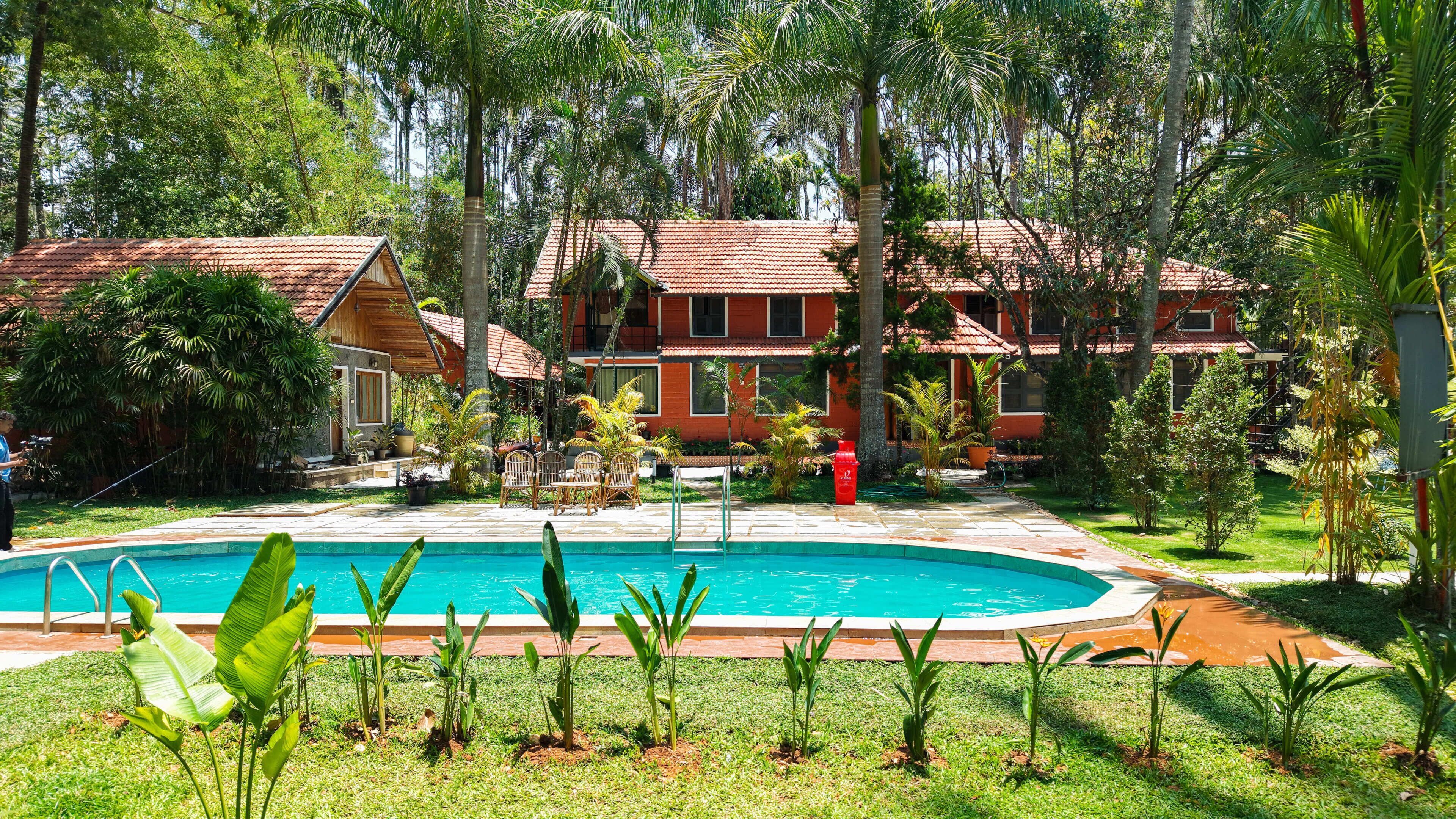 A pool surrounded by lush greenery, a wooden structure, and a red-roofed building.