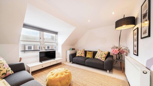 A modern loft apartment with a large skylight, a grey sofa with floral cushions, a yellow ottoman, and a floor lamp. - The Hideaway Cosy Seaside Getaway in Stonehaven (Stonehaven)