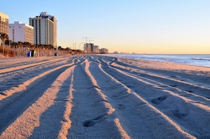 Ein Strand mit Fußspuren, die zum Meer führen, Gebäude im Hintergrund und einem klaren Himmel.