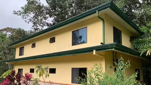 Un bâtiment à deux étages, doté d’une façade jaune et de bordures vertes, entouré d’une végétation luxuriante.