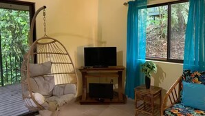 Une pièce avec un fauteuil suspendu, un téléviseur posé sur un meuble en bois, une petite table avec une plante et un canapé agrémenté d’un coussin à motifs floraux.