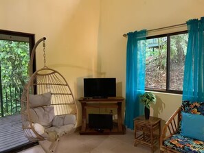 Ein Zimmer mit Hängekorbsessel, Fernseher auf einem Holzständer, kleinem Tisch mit Pflanze und Sofa mit Blumenkissen.