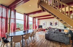 A spacious living area with a wooden floor, a dining table with chairs, a sofa, and a staircase.