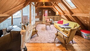 Et hyggeligt loftsværelse med træloft, en sofa med farverige puder, en ventilator og et vindue med udsigt over en gade.