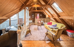 A cozy attic room with a wooden ceiling, a sofa with colorful cushions, a fan, and a window overlooking a street.