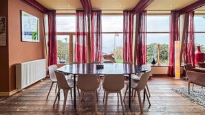 Un coin repas avec une table ronde et des chaises, de grandes fenêtres avec des rideaux, un radiateur et une vue sur la verdure à l’extérieur.