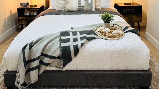 Un dormitorio con una cama grande, dos mesitas de noche, una mesita auxiliar y un cuadro enmarcado en la pared, además de una pequeña planta en maceta sobre la mesita.