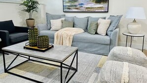 Un salón con un sofá gris, un sillón negro, una mesita de centro, una planta en maceta y un cuadro en la pared.