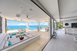 A modern living room with a large white sofa, a dining area with a bar, and a view of the ocean.
