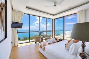 A spacious bedroom with a large bed, a desk, and a chair, all facing a large window with a view of the sea.