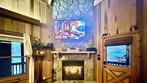 Una habitación acogedora con chimenea de piedra, paredes de madera, un televisor montado y vistas a través de una puerta.