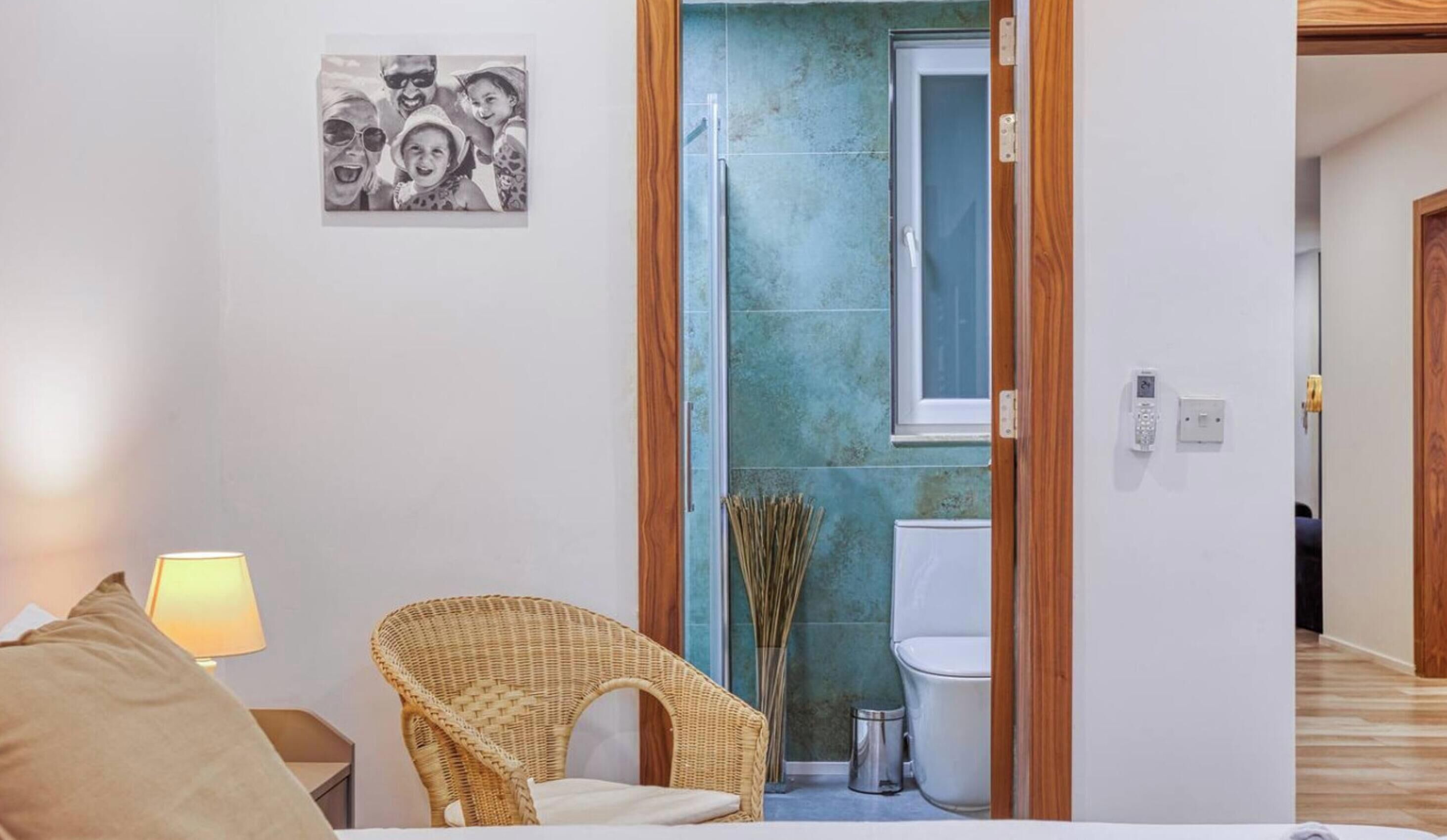 A room with a bed, a wicker chair, a lamp, a picture on the wall, and a bathroom with a toilet and shower.