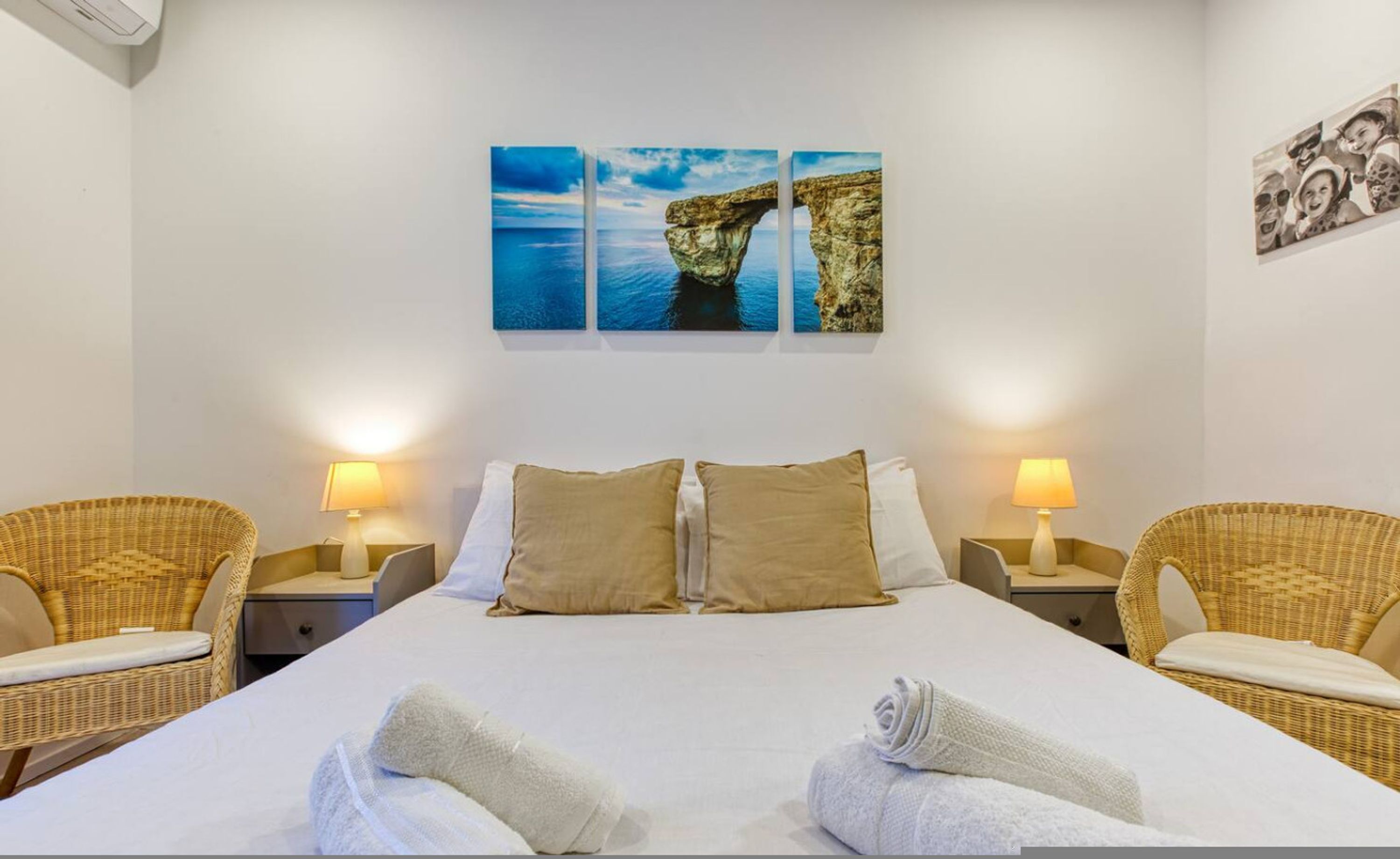 Una camera d'albergo con un letto, due comodini, due poltrone in vimini e un'opera d'arte a parete raffigurante una formazione rocciosa.