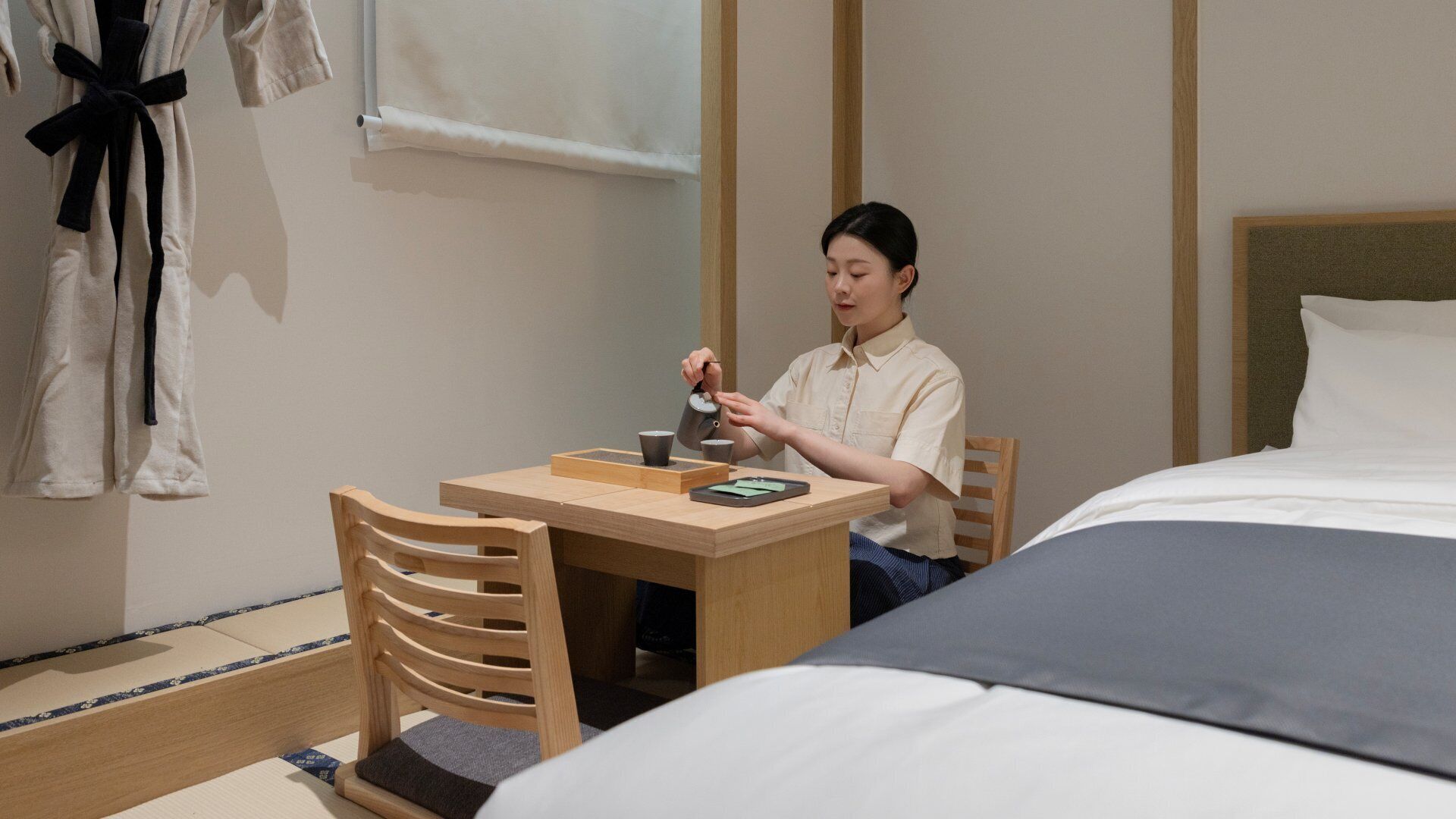 Una habitación de hotel con una cama, una silla de madera, una mesita y una persona sirviendo té.