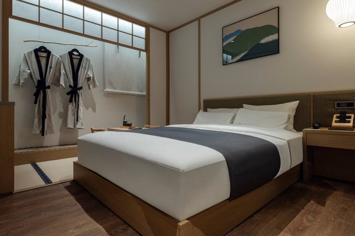 A hotel room with a bed, bedside table, nightstand, and a window with a view.