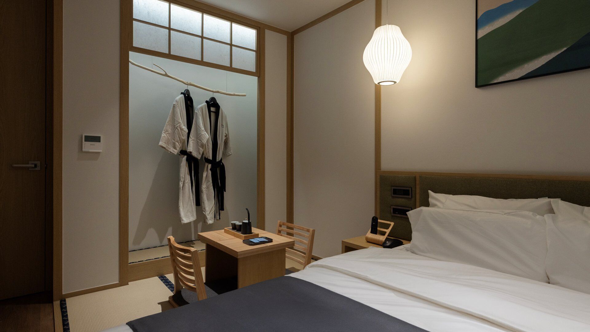 A hotel room with a bed, a desk with a chair, a robe rack, and a painting on the wall.