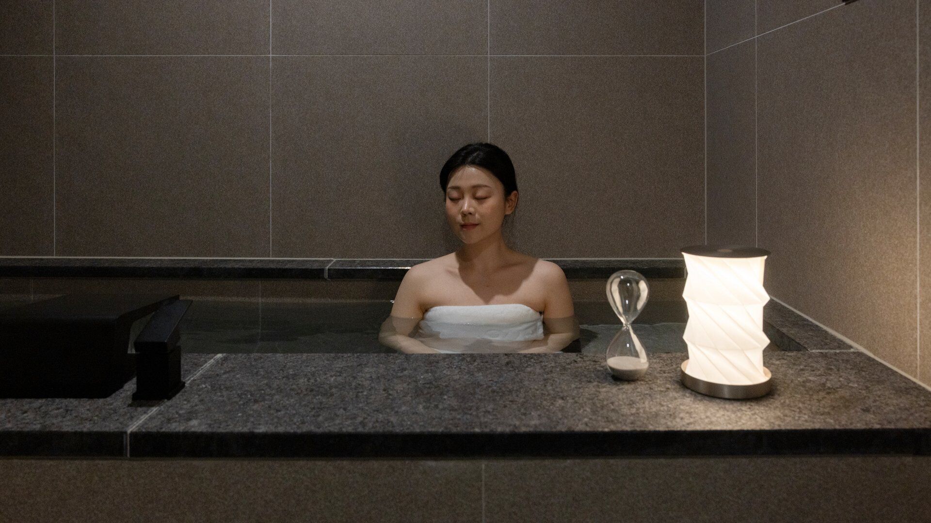 A person in a bathtub with a towel wrapped around their body, an hourglass, and a lamp on the side.