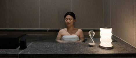 A person in a bathtub with a towel wrapped around their body, an hourglass, and a lamp on the side.