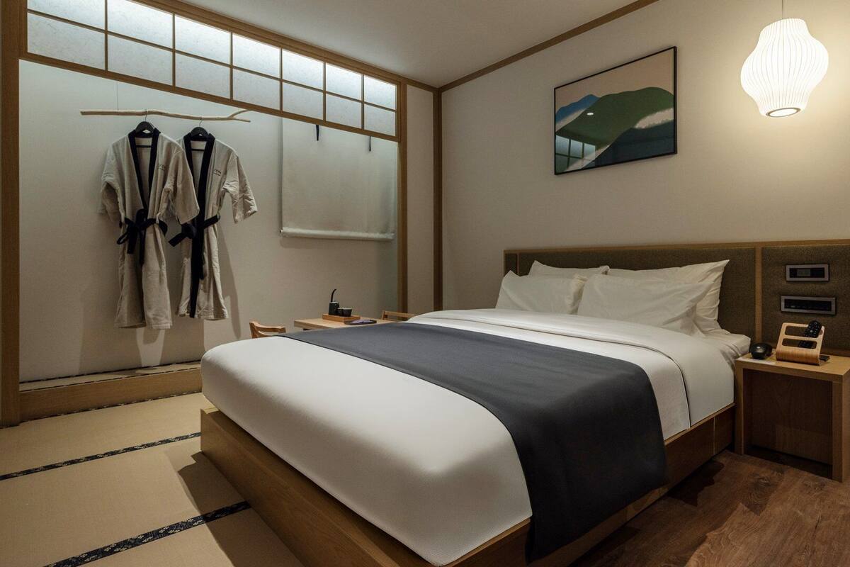 A hotel room with a bed, bedside tables, a nightstand with a lamp, a painting on the wall, and a window with a view.