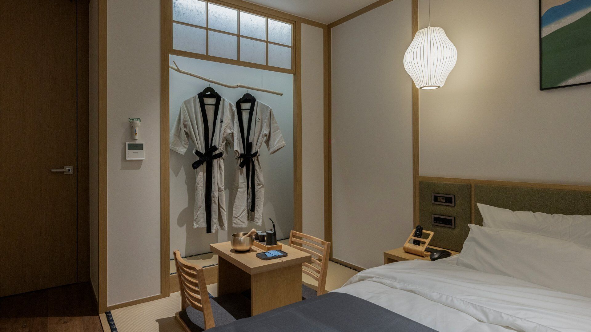 Et hotelværelse med seng, sengeborde, et skrivebord med stol, et knagered med to badekåber, et billede på væggen og en hængende lampe.
