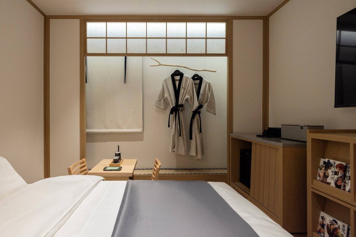 A hotel room with a bed, a small table with a cup and a book, a TV, and a wardrobe with pictures.