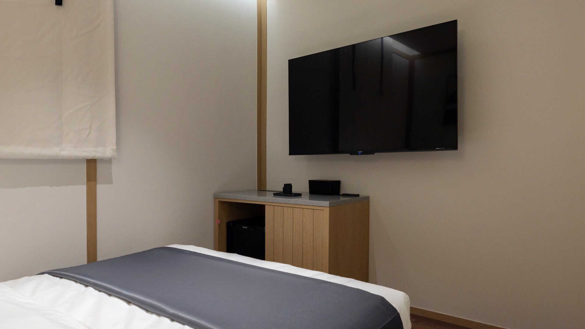 Hotellihuoneessa on sänky, pieni puinen yöpöytä, seinälle asennettu taulutelevisio ja ikkuna, jossa on valkoinen sälekaihdin.